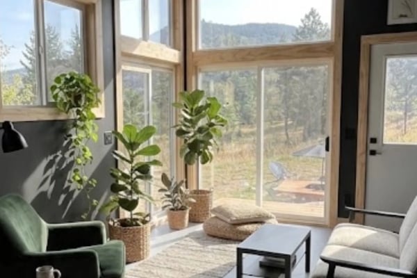 Modern living room with large windows overlooking forested mountains, potted plants on windowsill, green armchair and cream sofa.