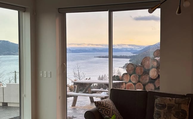 Living room with dark sofa and large windows overlooking a snow-covered lake surrounded by mountains at sunset
